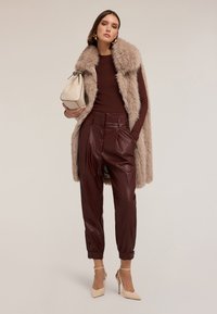 Kamizelka z sztucznego futra w odcieniu beżowym, brązowy top w prążki, bordowe skórzane spodnie z elastycznymi mankietami i beżowe szpilki; trzymając jasnobeżową torbę.