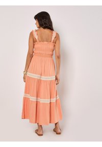 Koralli maksimekko, jossa on smockattu yläosa, röyhelömäiset olkaimet ja pitsikoristeet. Valoisa hame, jossa on kerroksia, valmistettu kevyistä materiaaleista.