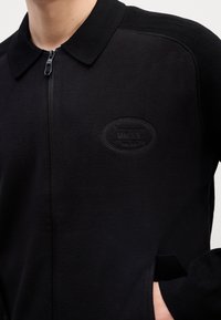 Veste noire zippée avec col, un logo ovale en relief "BOSS" sur la poitrine, et une main partiellement dans la poche latérale.