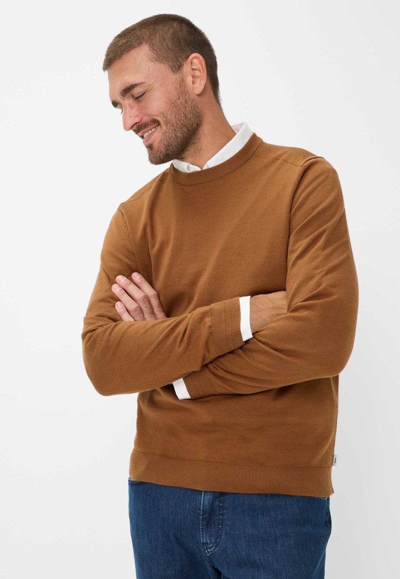 Bruine ronde hals trui met lange mouwen, geribde manchetten en zoom. Draagt over een witte blouse met kraag, gecombineerd met blauwe spijkerbroek.