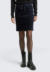 Jupe en velours côtelé bleu marine avec une taille à cordon, deux poches avant et une longueur genoux, associée à des bottines noires et des collants transparents.