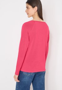 Femme aux longs cheveux bruns portant un haut à manches longues rose vif et un jean bleu, debout devant un fond blanc, légèrement tournée.