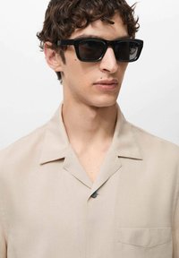 Beige kortebloemige shirt van gladde stof, met een notch-kraag, één pocket en een zwarte knop. Zwarte rechthoekige zonnebril.