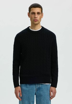 Homme portant un pull en maille torsadée noire par-dessus une chemise blanche et un jean bleu, debout devant un fond clair uni.
