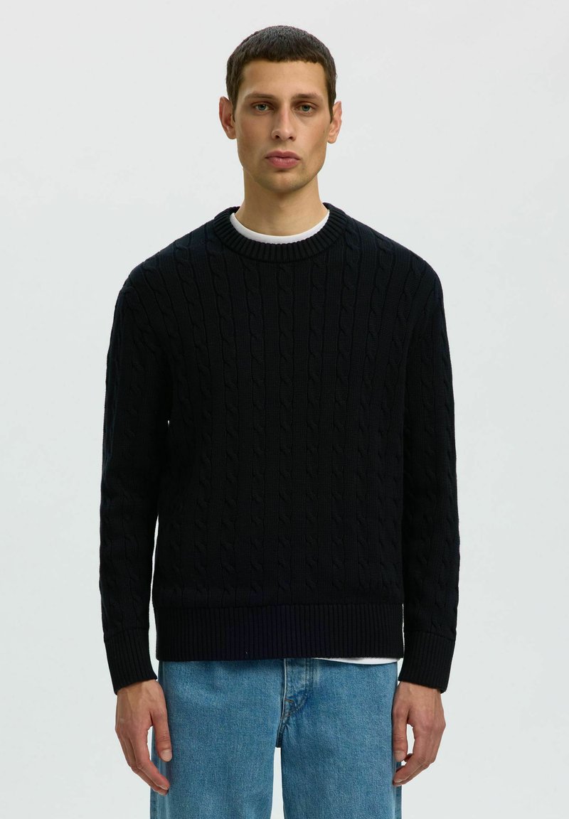 Homme portant un pull en maille torsadée noire par-dessus une chemise blanche et un jean bleu, debout devant un fond clair uni.