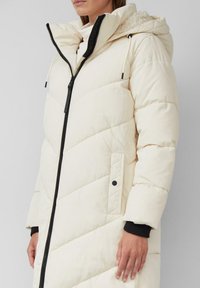 Gaišas krēms pufers jaka ar augstu apkakli, rāvējslēdzēja aizdari un melniem akcentiem. Iezīmēts ar kviltinga rakstu un elastīgām aprocēm siltumam.