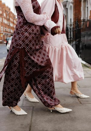 Two people walking side by side wearing patterned dresses and white low-heeled shoes with pearl details on a city sidewalk.