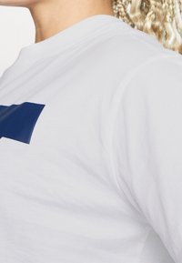 Valkoinen puuvillapaita, jossa on sininen logo edessä, pyöreä kaula-aukko ja lyhyet hihat, sileä materiaali ja rento istuvuus.