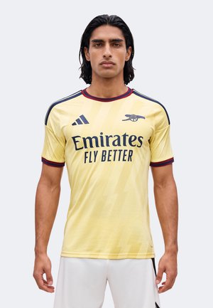 Uomo che indossa la maglia da calcio gialla dell'Arsenal con il logo dello sponsor "Emirates Fly Better", in piedi davanti a uno sfondo chiaro uniforme.