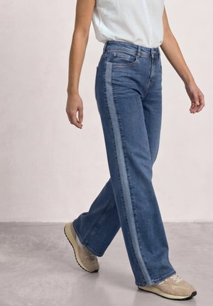 Person, der går iført blå jeans med en lysere sidelstribe, en hvid ærmeløs skjorte og beige sneakers på en grå gulv.