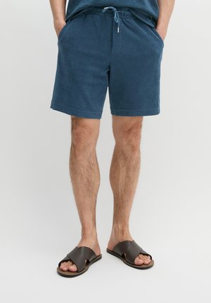 Man draagt blauwe shorts met trekkoord en bruine sandalen met kruisbandjes, staat met handen in de zakken tegen een effen achtergrond.