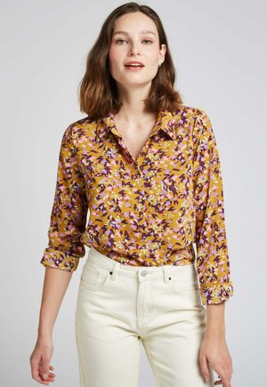 Chemisier moutarde avec un motif floral violet et rose, col boutonné, manches longues, associé à un jean blanc taille haute.