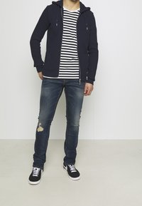 Námořnicky modrá mikina s kapucí na zip a přední kapsou, pruhované bílo-černé tričko, modré džíny s efektem opotřebení a černé tenisky s bílou podrážkou.