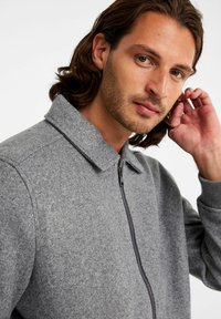 Grijze zip-up jas van textuurstof met een kraag en zichtbare stiksels langs de voorkant. Soepele ritsdetails.