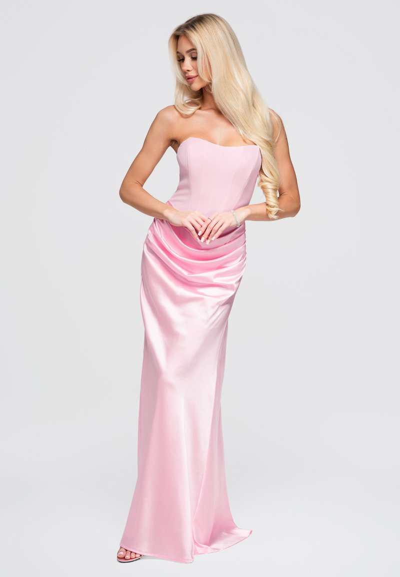 Femme blonde portant une robe en satin rose pâle sans bretelles, debout, les mains jointes, regardant vers le bas sur le côté sur un fond blanc uni.