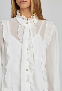 Weiße Bluse aus transparenter Stoff, mit Rüschen entlang der Vorderseite und des Kragens, einem gebundenen Halsdetail und einem dekorativen Knopf.