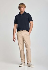 Marinblå piké med korta ärmar och en röd logotyp, kombinerad med ljusbeiga chinos och grå sneakers, stående mot en neutral bakgrund.