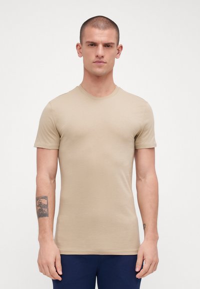 Beige short-sleeve T-shirt, round neck, fitted design, soft texture, smooth fabric, worn with dark blue pants, with a tattoo on the left forearm.
