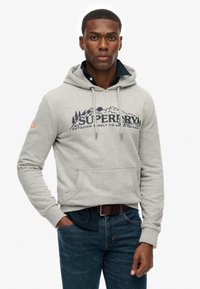 Grijze hoodie met een grote "SUPERDRY"-logo met berg- en wolkgraphics, kangoeroezak en geribbelde boorden. Gecombineerd met een blauwe spijkerbroek.