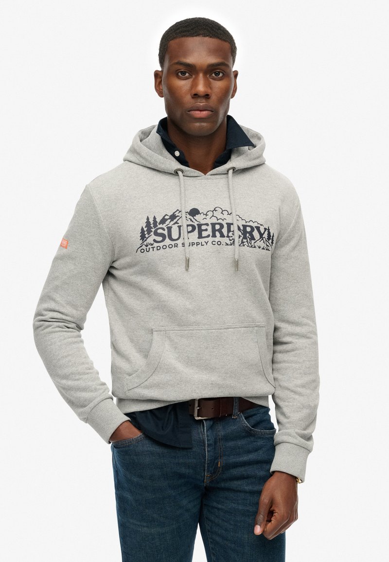 Grijze hoodie met een grote "SUPERDRY"-logo met berg- en wolkgraphics, kangoeroezak en geribbelde boorden. Gecombineerd met een blauwe spijkerbroek.