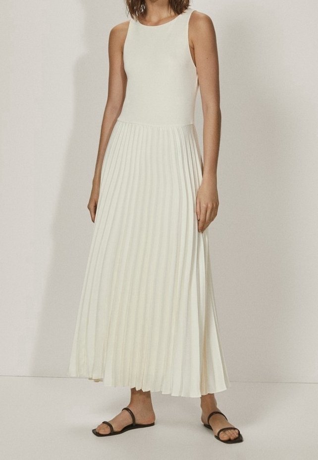 Femme portant une robe blanche sans manches avec une jupe plissée et des sandales plates noires, debout devant un fond uni.