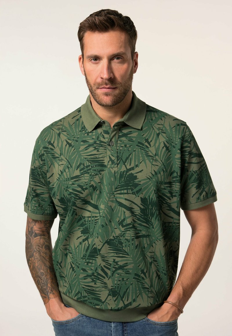 Grünes Poloshirt mit einem Blattmuster, kurzen Ärmeln und einem Kragen. Das Textil wirkt weich mit einem leichten Schimmer. Wird mit blauen Jeans getragen.
