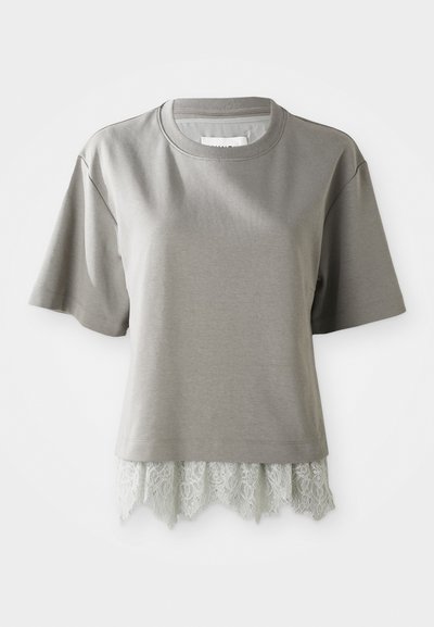 Kortermet, lys grå T-skjorte i mykt materiale, med blondekant nederst med bølget kant. Strukturert blonde gir detalj.