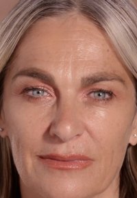 Close-up van een gezicht met een gladde huid, blauwe ogen, lichtbruin haar en minimale make-up. Opvallende kenmerken zijn fijne lijntjes en een neutrale lipkleur.