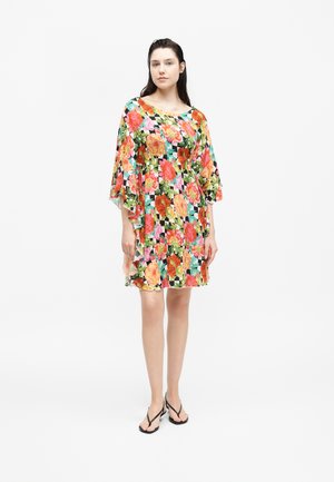 Vrouw draagt een kleurrijke jurk met bloemenprint, losse wijde mouwen en zwarte teenslippers, staand tegen een effen witte achtergrond.