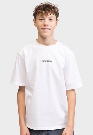 Έφηβος με σγουρά μαλλιά που φοράει λευκό T-shirt με τη φράση "HOUSE RULES" και μπλε τζιν, στέκεται μπροστά σε ανοιχτό γκρι φόντο.