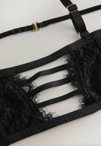 Juodas nėrinių liemenukas, turintis kelias elastingas juostas, nėrinių apdailą ir reguliuojamas satino juostas su auksinės spalvos detalių akcentais.