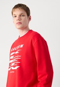 Sarkana pūkste ar baltu grafiku, kurā ir teksts "CALVIN" un stilizētas līnijas. Gluda auduma, ar apaļu kakla izgriezumu, brīvs piegriezums.