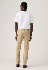 Χακί παντελόνι με στενή εφαρμογή, με πίσω τσέπες και κόκκινο λογότυπο. Συνδυασμένο με απλό λευκό T-shirt και σκούρα παπούτσια.