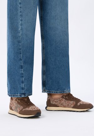 Person trägt blaue Jeans und braune Sneakers mit strukturiertem Stoff und beigen Sohlen und steht auf einer weißen Oberfläche.