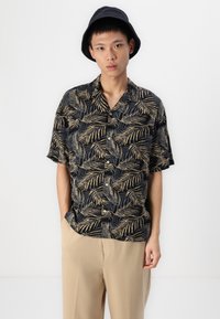 Camisa de manga corta en negro con un patrón de hojas tropicales doradas, con botones y un cuello clásico. Combinada con pantalones beige.