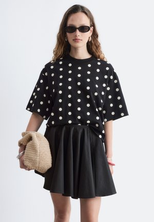 Femme portant des lunettes de soleil noires, un t-shirt noir à pois blancs, une jupe plissée noire, tenant une pochette tricotée beige sur fond uni.