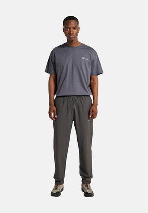 Ein Mann steht nach vorne gerichtet und trägt ein dunkelgraues T-Shirt sowie dazu passende dunkelgraue Hosen mit elastischem Bund und beigen Wanderschuhen.