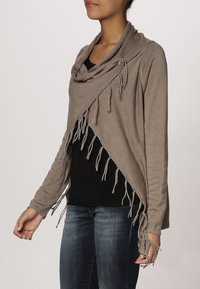 Wrap grigio a maglia con colletto drappeggiato, dettagli in frange all'orlo e bottoni decorativi. Indossato sopra una maglietta nera e jeans blu.