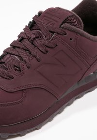 Baskets New Balance 574 bordeaux avec lacets texturés, col rembourré, dessus en suède et mesh, et logo embossé sur le côté.