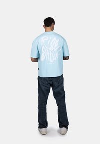 Světle modré oversized tričko s bílým grafickým textem "RISE & SHINE" na zádech, spárované s tmavě modrými džíny a bílými teniskami.