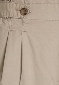 Beige katoenen stof met een gestructureerde afwerking, voorzien van een enkele bruine knop en een plooidetail, netjes gestikt langs de tailleband.