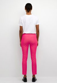 Pantaloni aderenti rosa brillante con una linea dritta, due tasche posteriori e una leggera texture, abbinati a una t-shirt bianca semplice.
