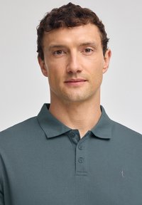 Dunkelblau-grünes Poloshirt mit weichem Material, dreiknöpfigem Schlitz und kleinem gesticktem Logo auf der Brust.