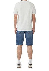 Witte katoenen t-shirt met korte mouwen, gecombineerd met blauwe denim shorts. Draagt witte sokken en schoenen met zwarte accenten.