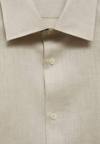 Camicia di lino beige con colletto a punta, caratterizzata da una fila di due bottoni sulla patta, e un tessuto lavorato e texturizzato.