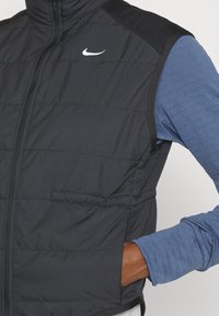 Sort polstret vest med lynlåslukning, der har et hvidt Nike-logo på brystet og en sort panel ved skuldrene. Bær over en blå langærmet skjorte.