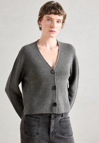 Cardigan cinza pregueado com decote em V, apresentando três botões pretos, mangas longas e um design cropped. Usado sobre uma saia de ganga cinza.