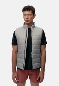 Lys grå polstret vest med ståkrage, frontglidelås og quiltet tekstur. Brukt over en svart t-skjorte og rosa shorts.