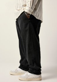 Pantalones de mezclilla negros con un corte relajado, combinados con zapatillas blancas y grises. La camisa es color crema con rayas verticales negras.