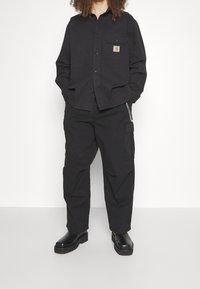 Mustat paidat, joissa on nappikiinnitys, yksi rintatasku ja logopatch. Yhdistetty mustiin cargo-housuihin ja massiivisiin mustiin saappaisiin, kaikki puuvillakankaasta.